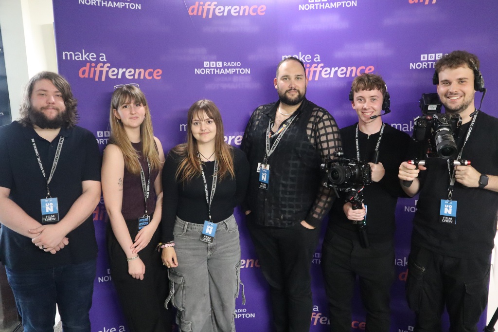 Six people wearing lanyards stand in front of a purple "Make a Difference" BBC Radio Northampton backdrop, with two holding professional cameras.