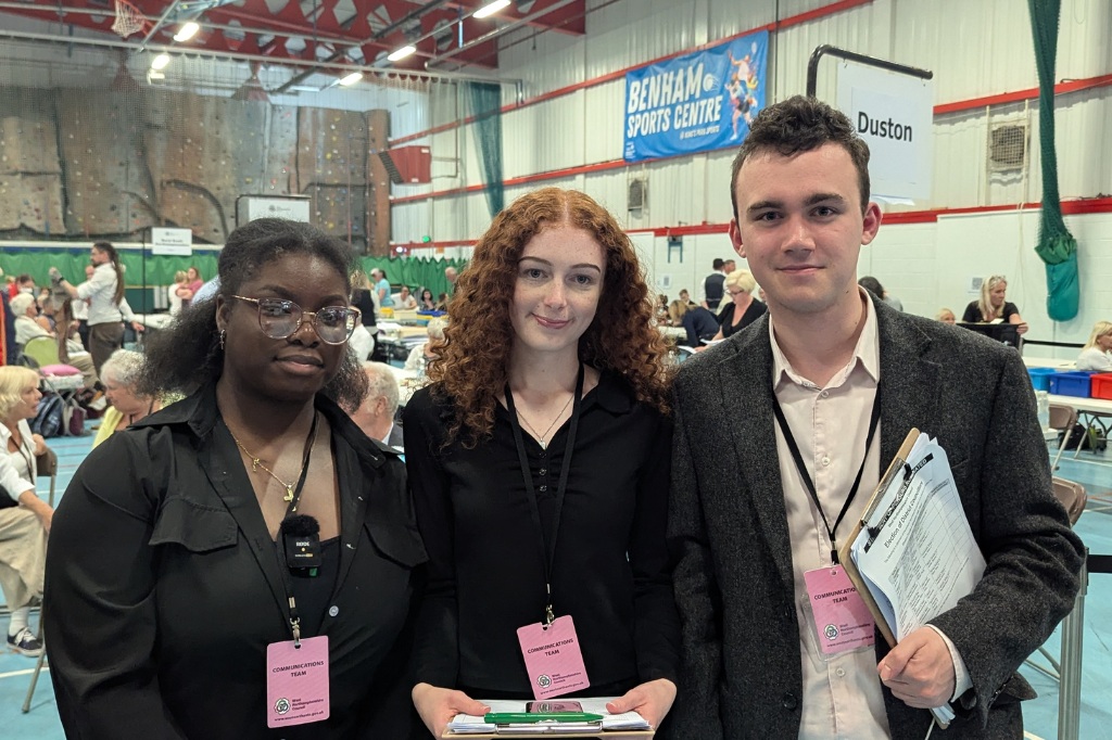 Three young people with name badges and clipboards stand indoors at an event in a sports hall, with tables and other attendees visible in the background.