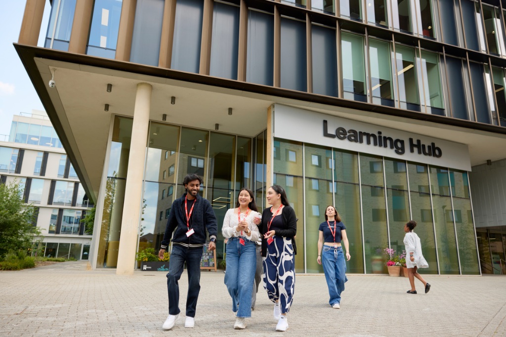 Four students walk and talk outside a modern building labeled "Learning Hub" on a university campus.