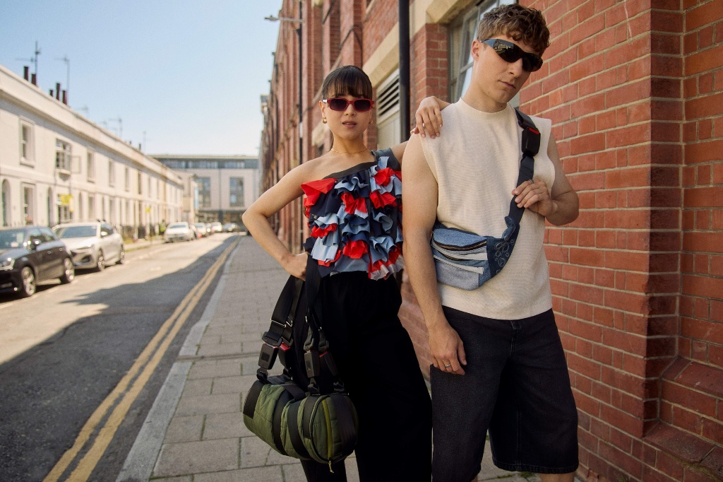 Two people stand on a city sidewalk, both wearing sunglasses and carrying bags. One wears a ruffled top and black pants; the other wears a sleeveless shirt and denim shorts.