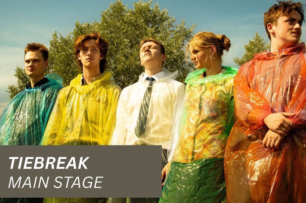 Five young adults stand outdoors; four wear colorful rain ponchos and one in a shirt and tie. Overlay text reads: "TIEBREAK MAIN STAGE." Trees and blue sky are in the background.