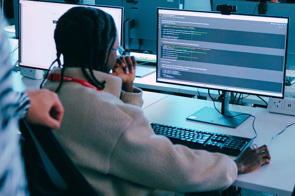 A person sits at a desk, looking at code on a computer monitor in an office setting, with another person standing nearby.