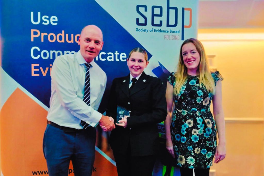 Three people pose for a photo; one shakes hands with another who holds a glass award, standing in front of a Society of Evidence Based Policing banner.