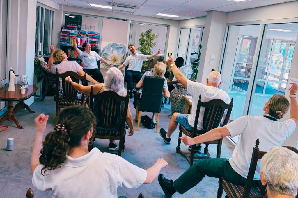 A group of older adults seated in chairs participate in a guided exercise session in a bright, indoor room with large windows.