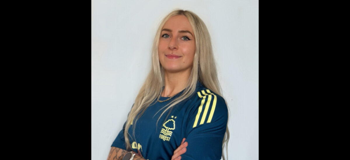 Briony Garner, a Sports Rehabilitation and Conditioning BSc graduate stands against a plain background. She has long blonde hair and visible arm tattoos, and is standing wearing a blue Nottingham Forest football jersey with yellow accents, arms crossed.