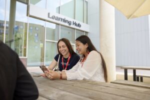 Two women wearing red lanyards sit at an outdoor table, looking at a phone and smiling, with a building labeled "Learning Hub" in the background.