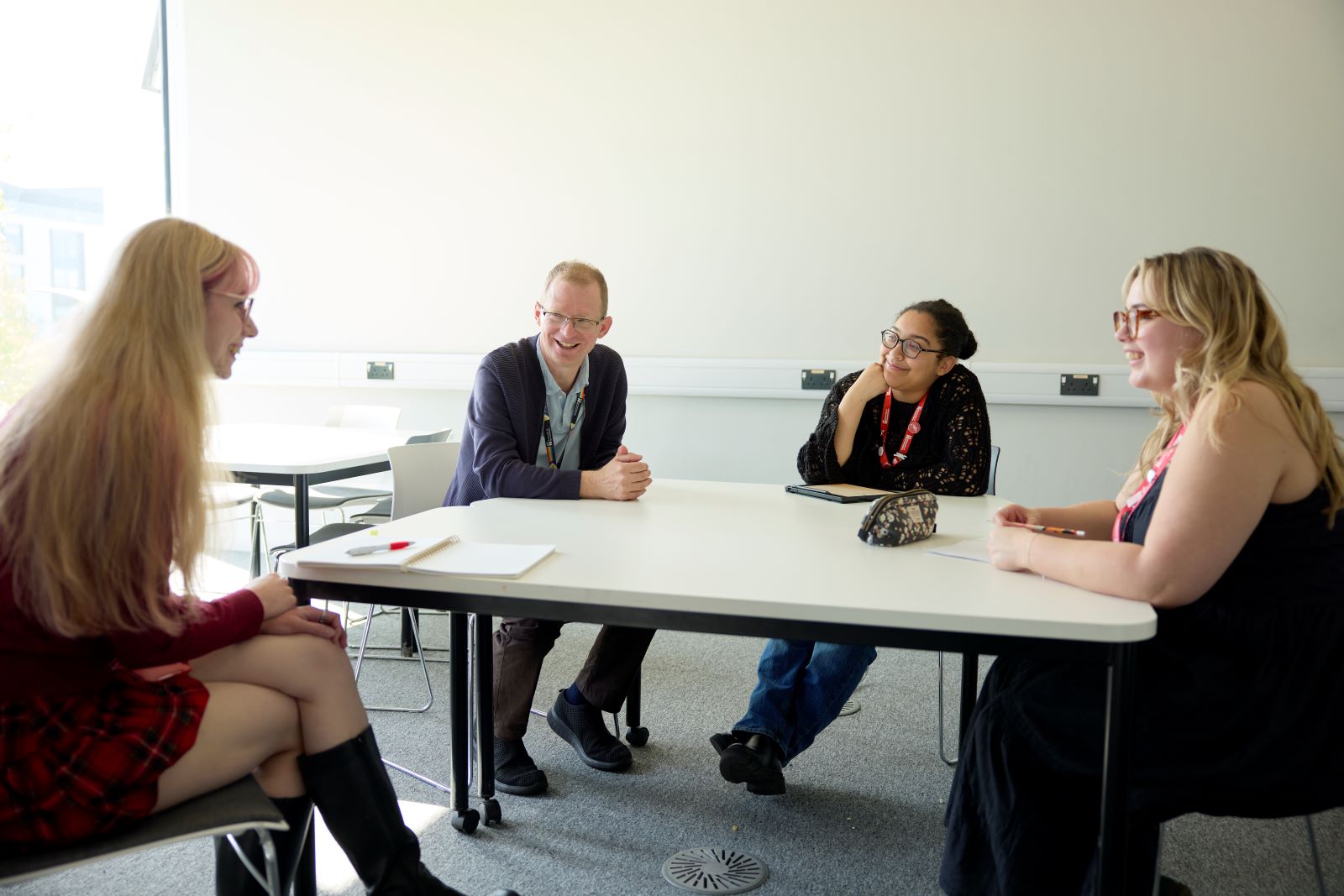An academic and three students sit around a table in a well-lit room, engaged in conversation. There are notebooks and a pencil case on the table.