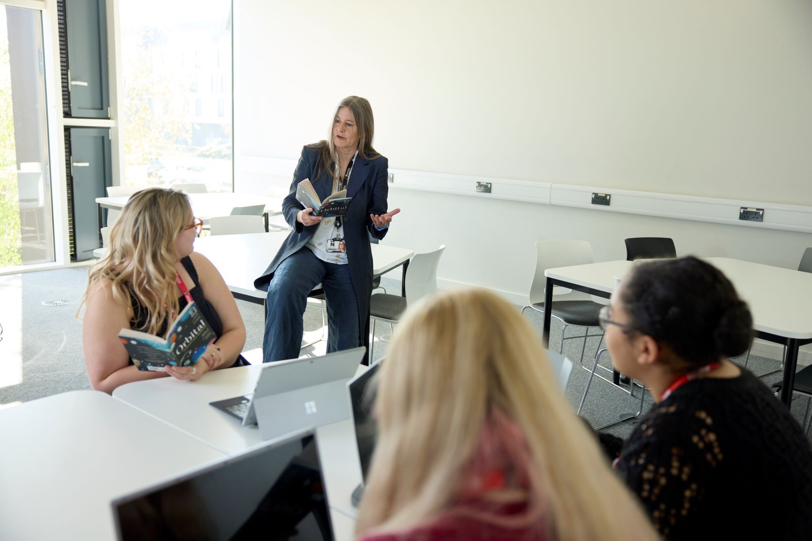 An academic leans on a table and speaks to three seated students in a classroom. All are holding books and laptops are on the table. The room has white walls and large windows.