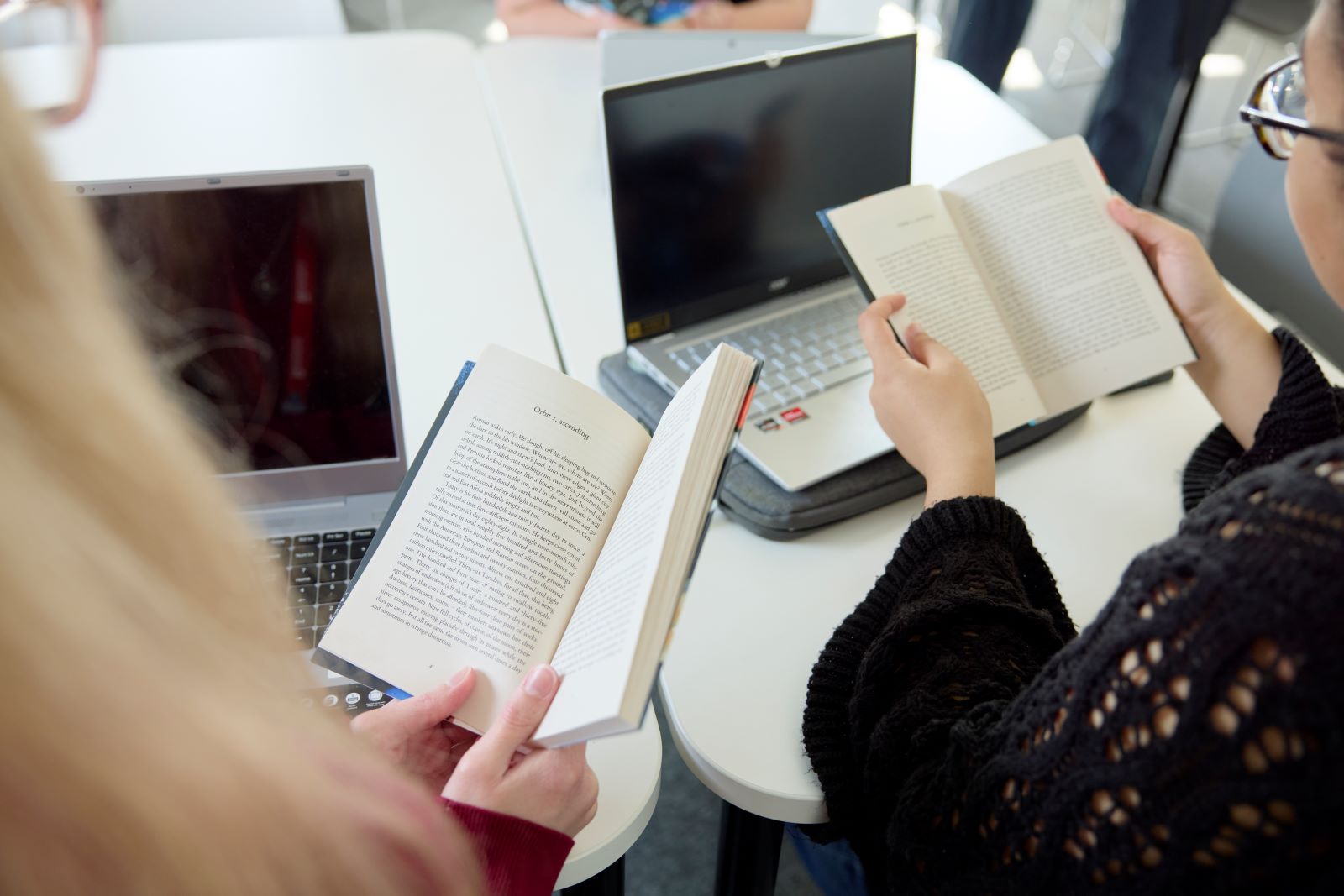 Two people sit at a table with open laptops, each holding and reading a book.