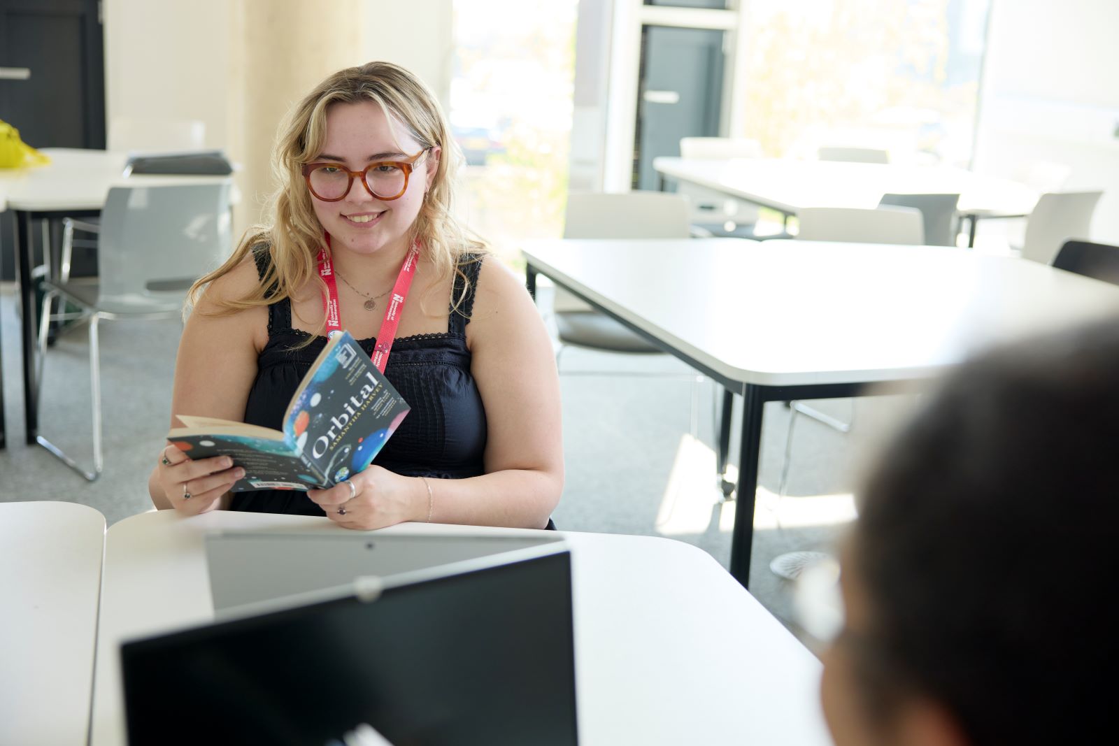 A student wearing glasses and a lanyard sits at a table holding a book, smiling at someone across from her in a bright, modern room with empty chairs and tables.