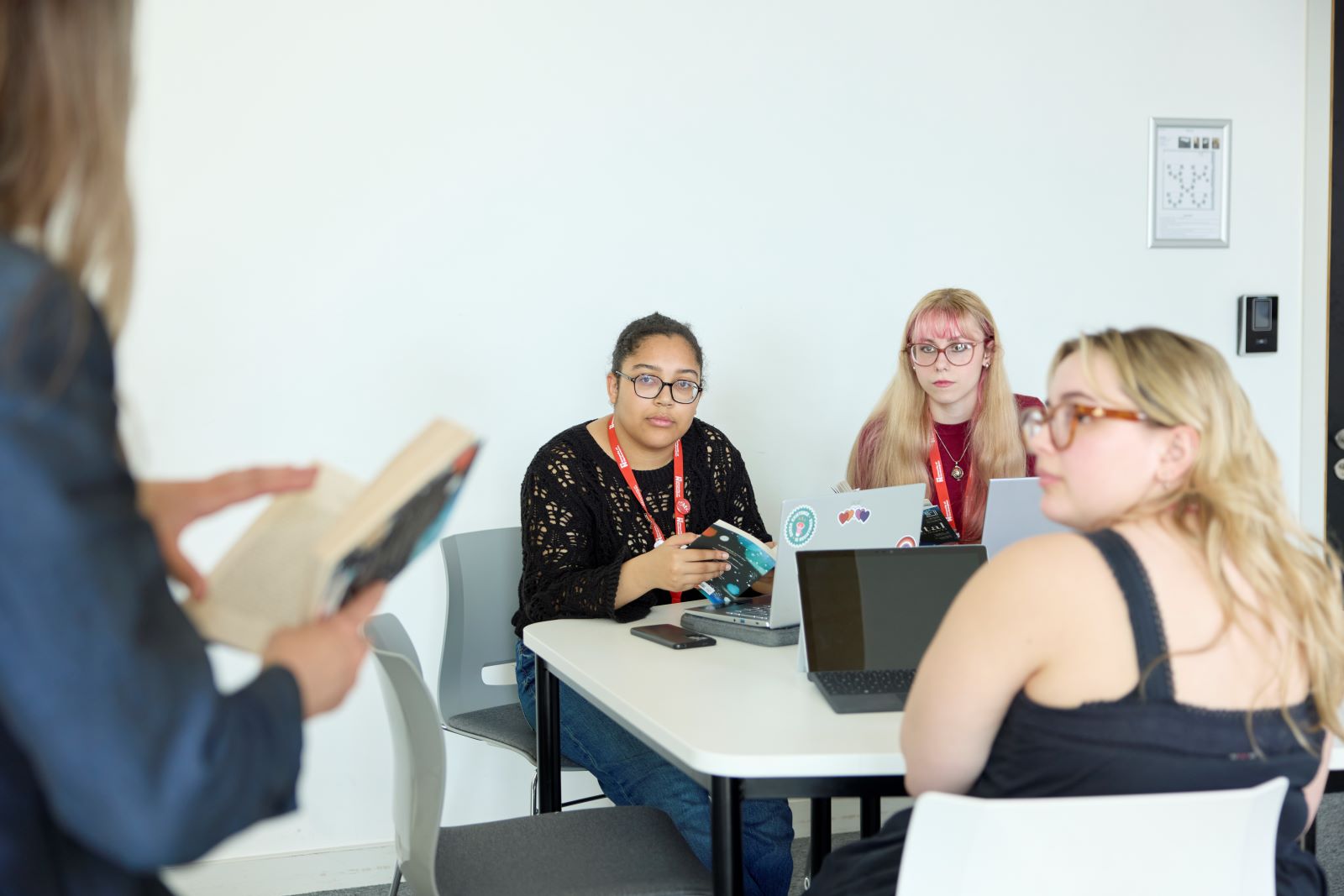 Three students sit at a table with laptops, listening to an an academic standing nearby holding a book in a bright, modern room.