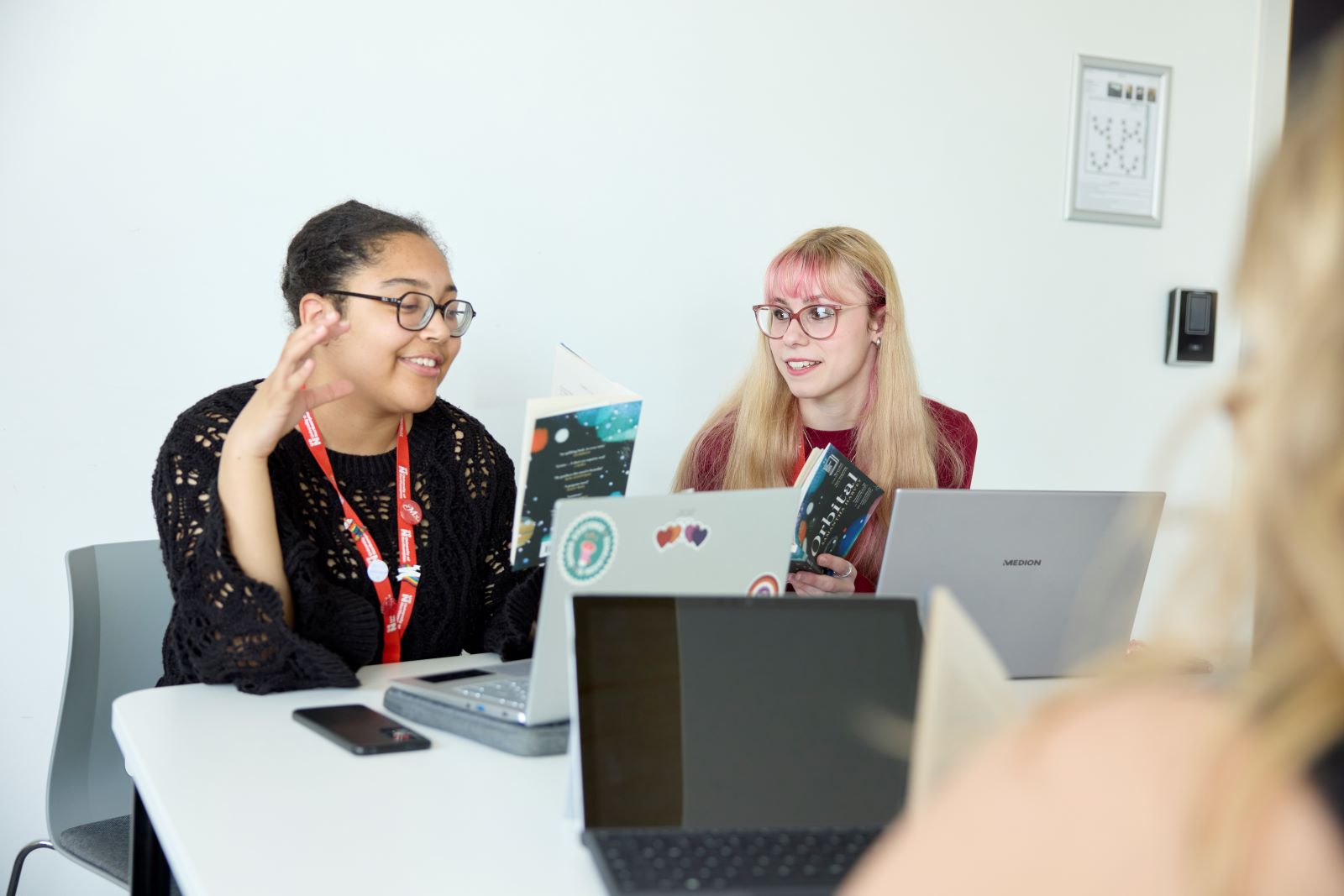 Two people sit at a table with laptops and books, engaged in conversation in a bright room. A third person is partially visible in the foreground.