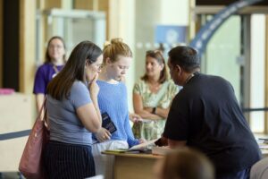 Two visitors stand at a registration counter talking to a staff member at a UON Open Evening, while two others look on in the background in a brightly lit indoor setting.