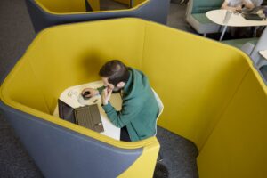 A person sits at a desk with a laptop in a yellow pod, working and using a mouse.