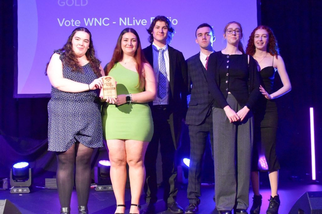 Six people dressed in formal attire stand on a stage, with one person holding a wooden award. A purple-lit screen behind them reads, "Vote WNC - NLive Radio.
