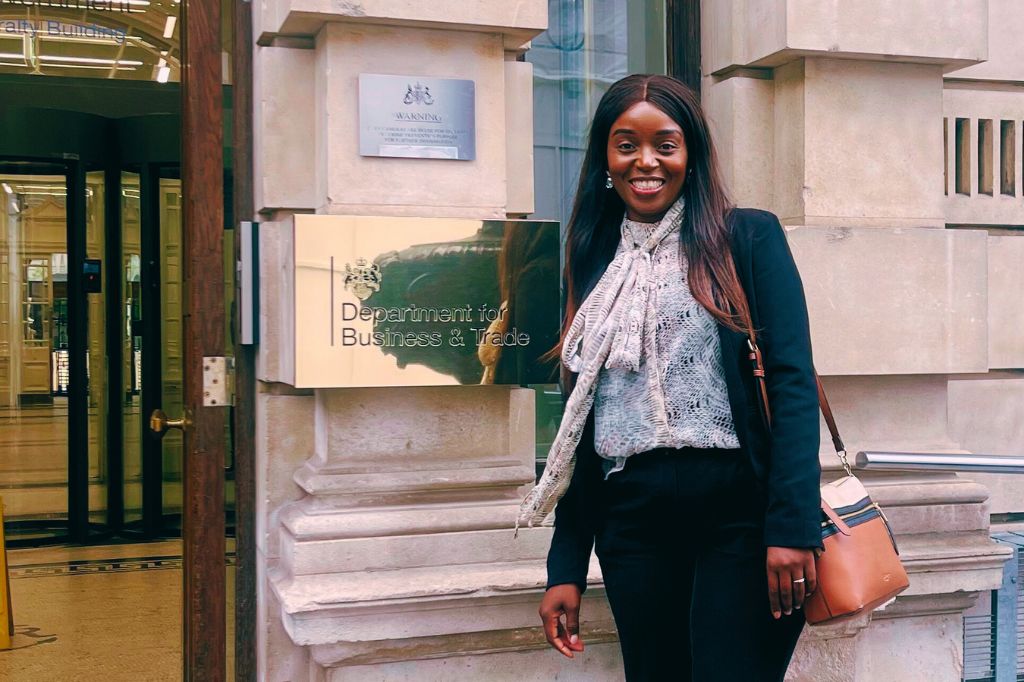 A woman stands smiling in front of a building entrance, next to a sign that reads 'Department for Business & Trade.'