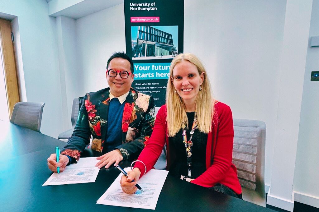 Two people sit at a table, smiling and signing documents, with a University of Northampton banner in the background.