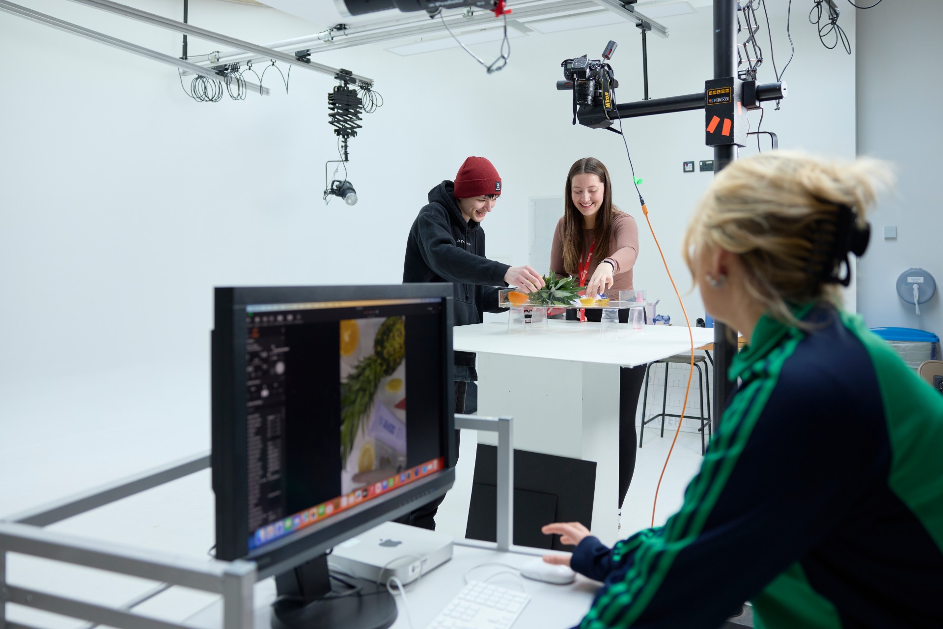 One student sat at a screen viewing the camera imagery, whilst two students adjust and move products such as a plant and clear cups into position.
