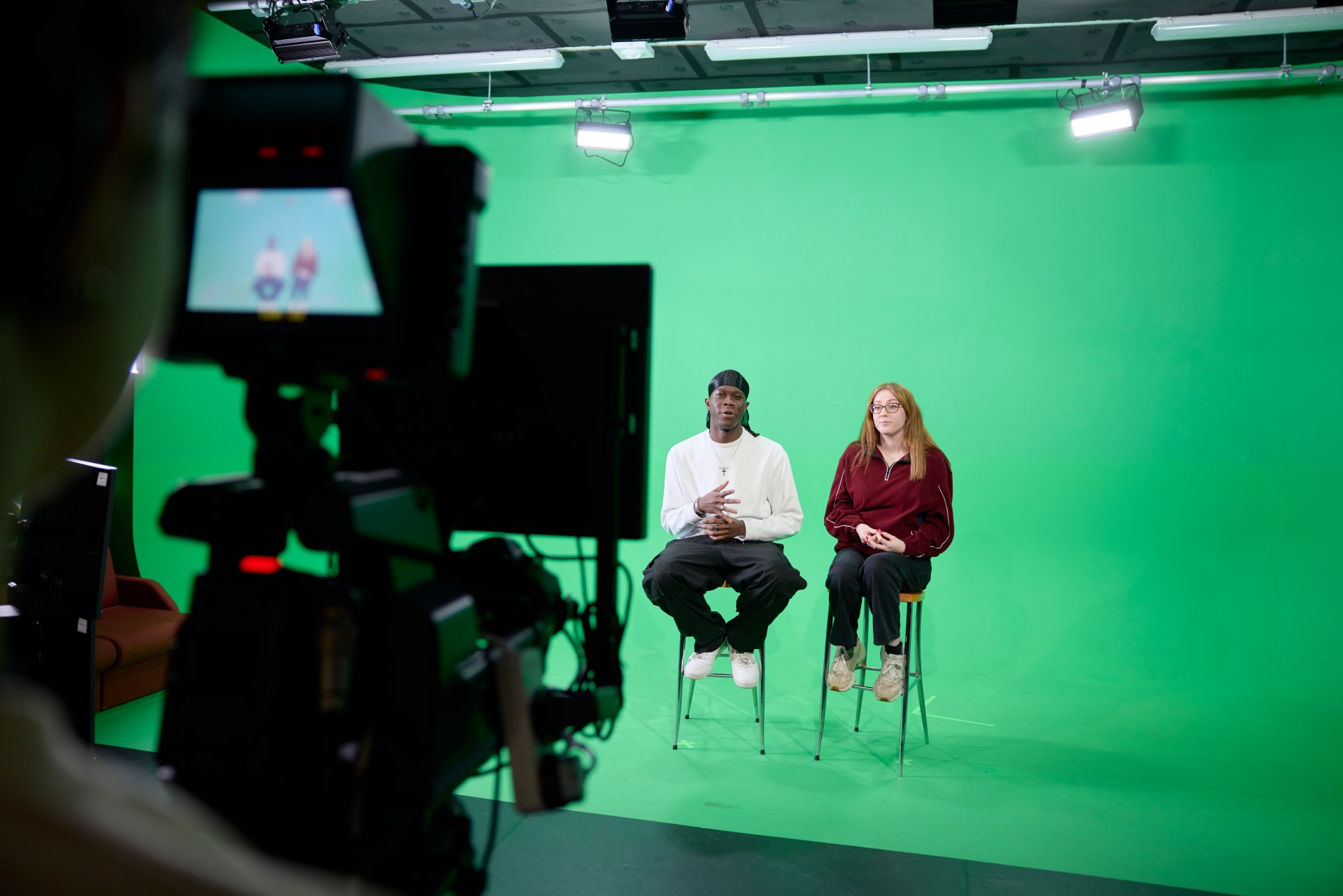 Two students seated on stools in front of a green screen, preparing for a TV studio recording during a university journalism workshop, with a camera in the foreground