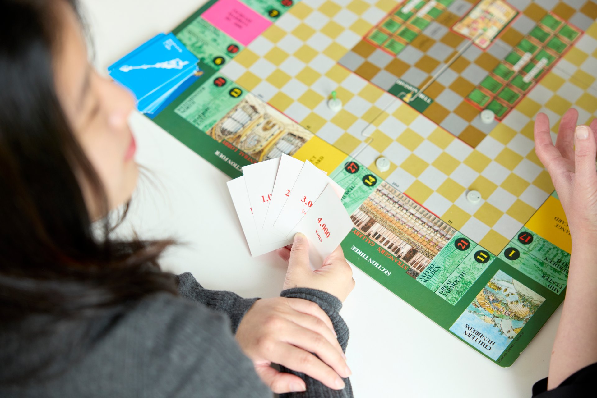 Two university students playing a strategy board game featuring cards and a colorful checkered board with illustrations of buildings and landmarks