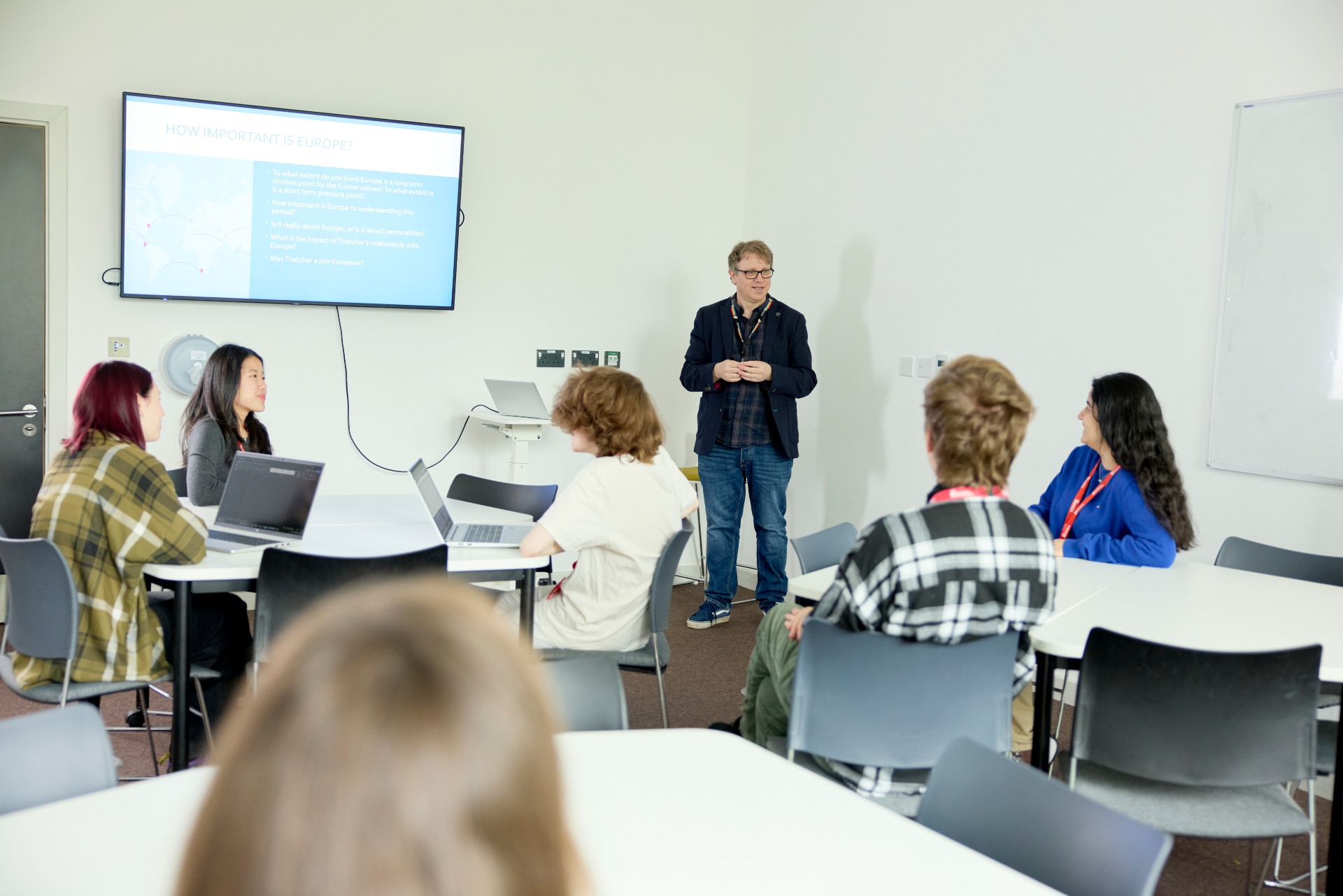 A lecturer stands at the front of a classroom giving a presentation titled “How Important is Europe?” to a group of students seated around tables with laptops, engaged in discussion at the University of Northampton.