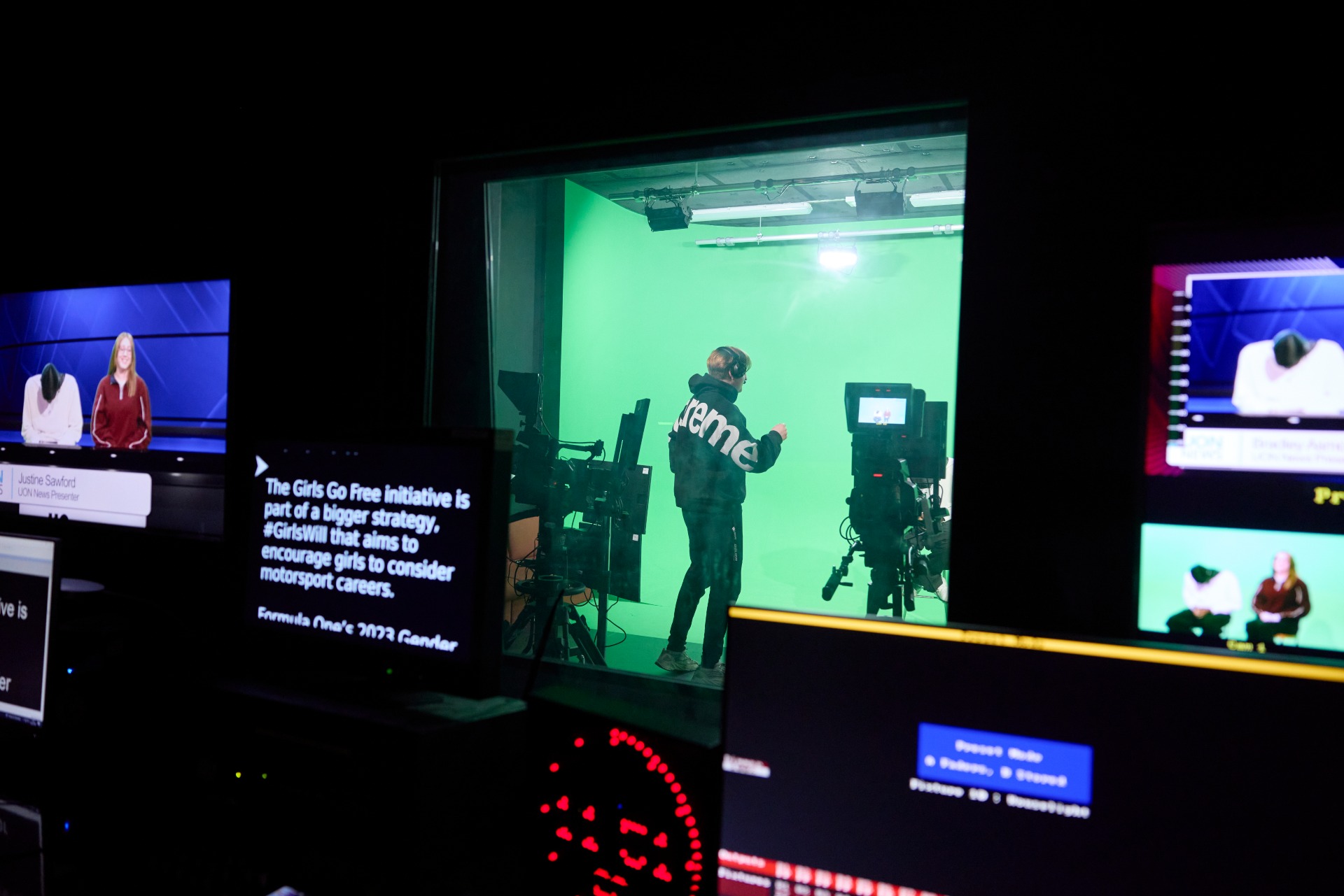 View from the control room into the TV studio, showing a student operating a camera in front of a green screen while multiple monitors display live feeds