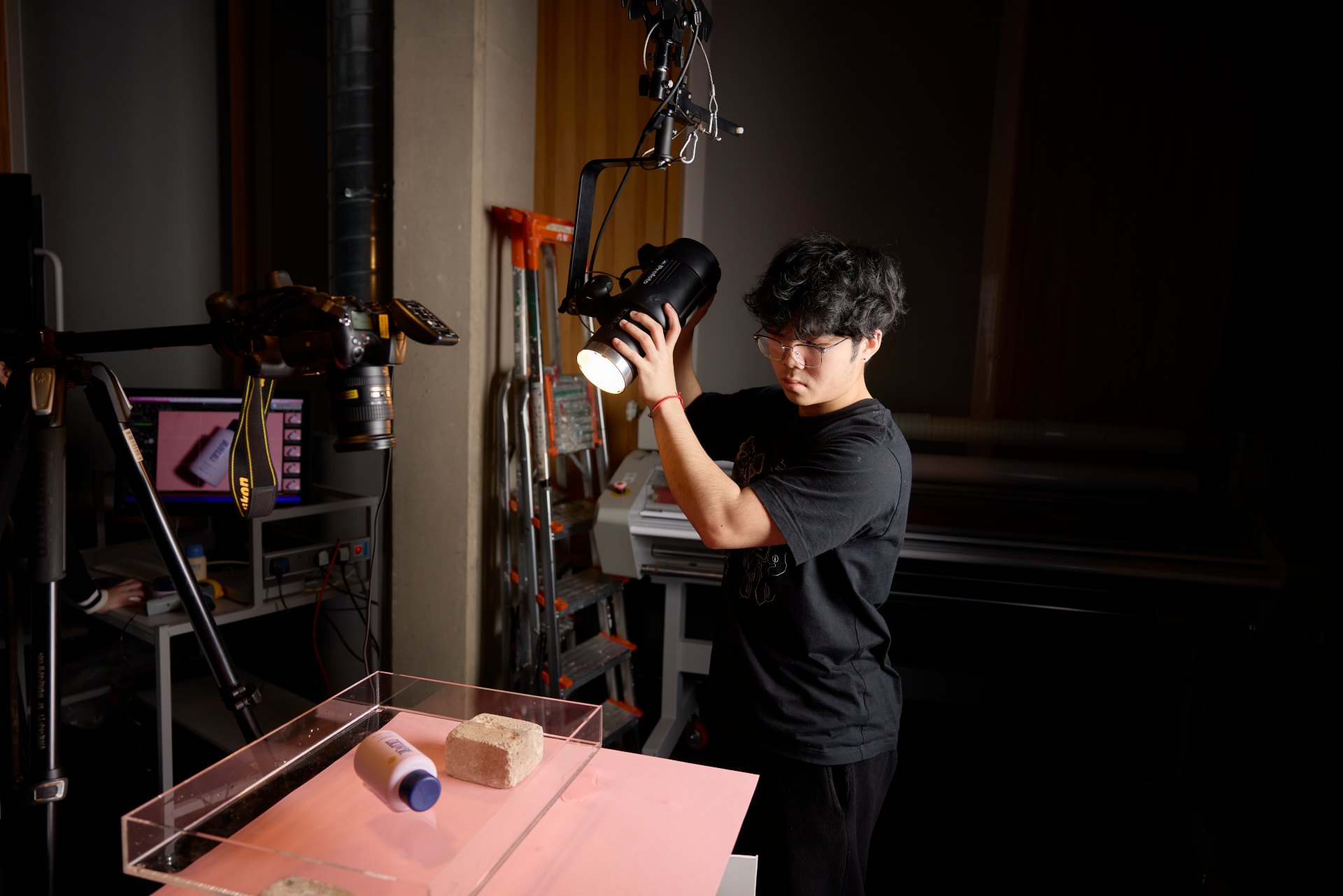 Student adjusting a handheld studio light over a product setup on a pink surface during a photography workshop, with a mounted camera and computer workstation visible in the background