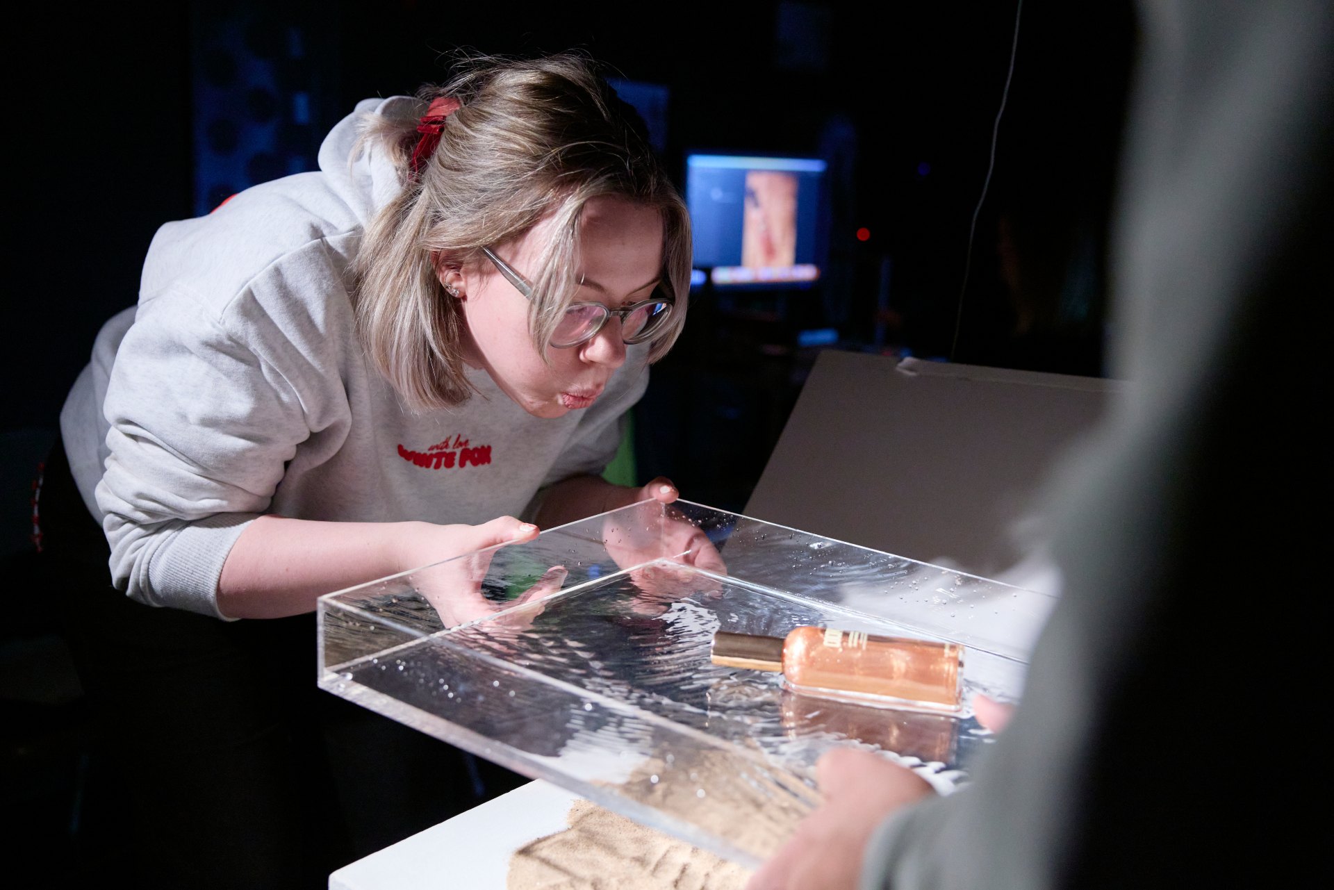Student in a photography studio adjusting a transparent surface with a bottle on a clear background, as part of a product photography setup during a university workshop