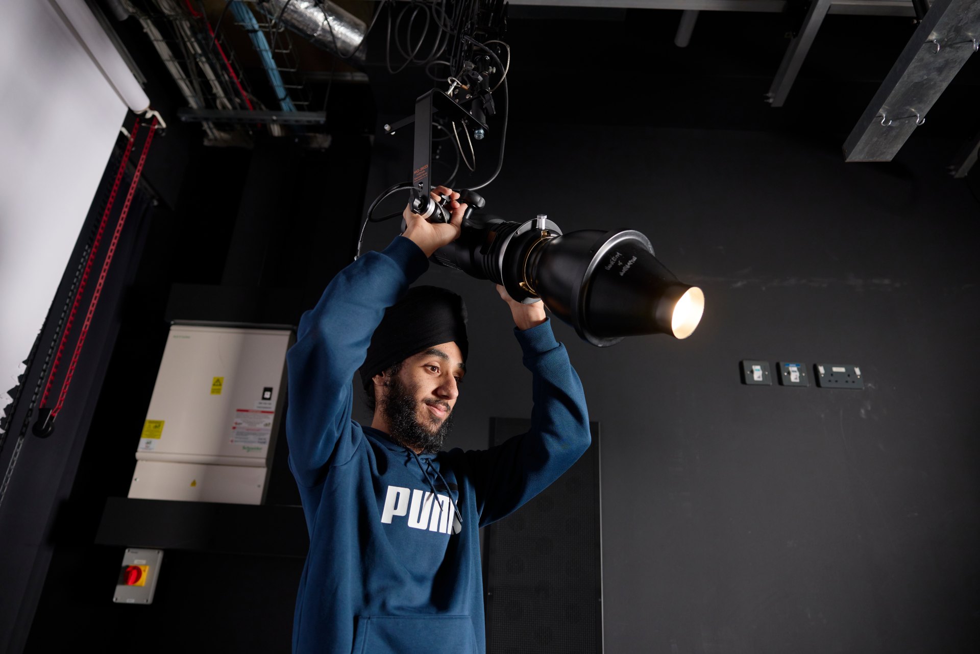 Student adjusting a large studio light mounted on an overhead rig in a professional photography studio during a lighting setup exercise