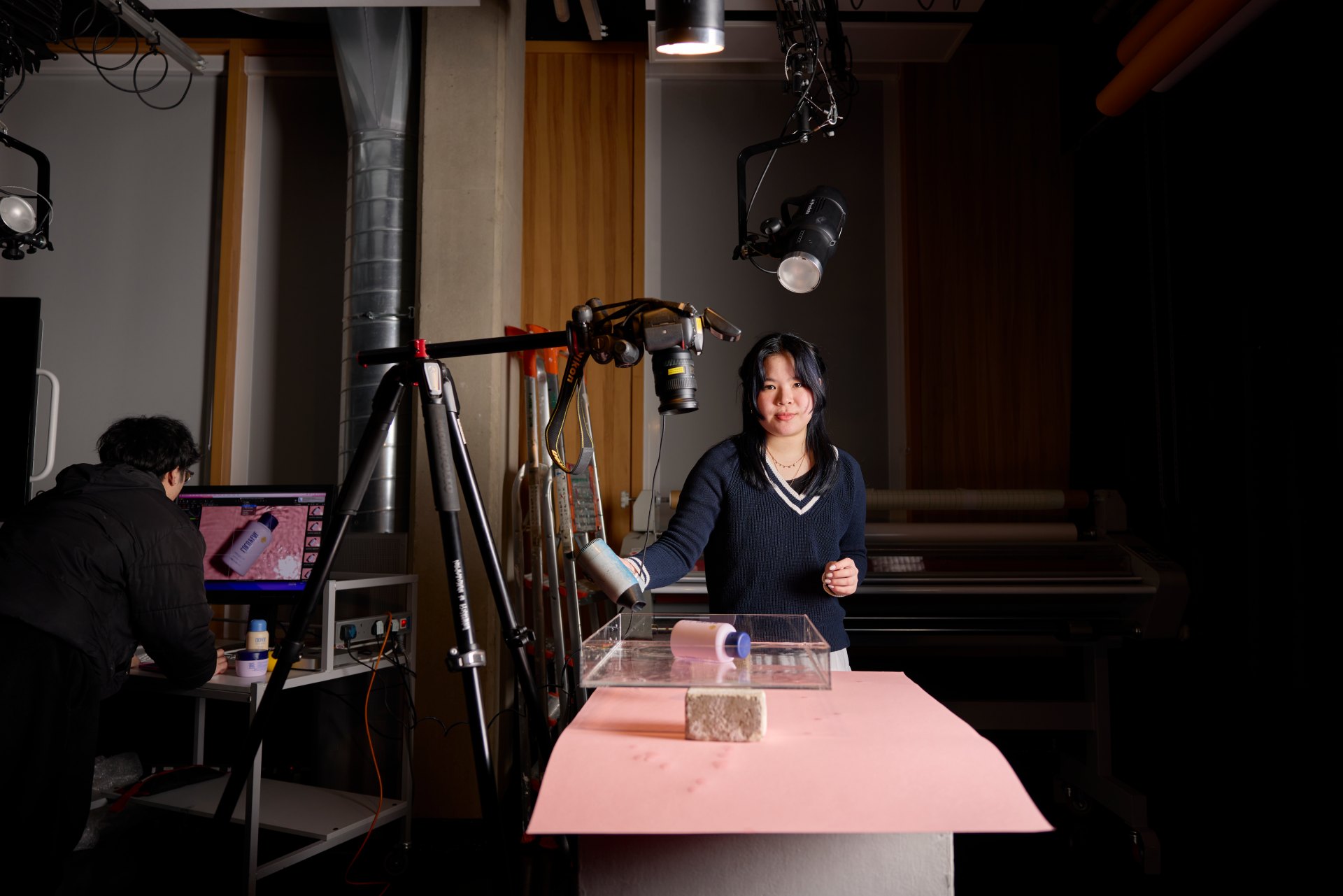 Students working in a university photography studio, with one arranging an object on a pink surface under a mounted camera while another monitors the setup on a computer screen