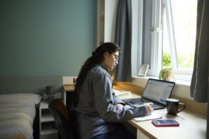 A student at a desk inside student halls writing with a pen in their right hand, a notepad on the desk, and a laptop in front of them.