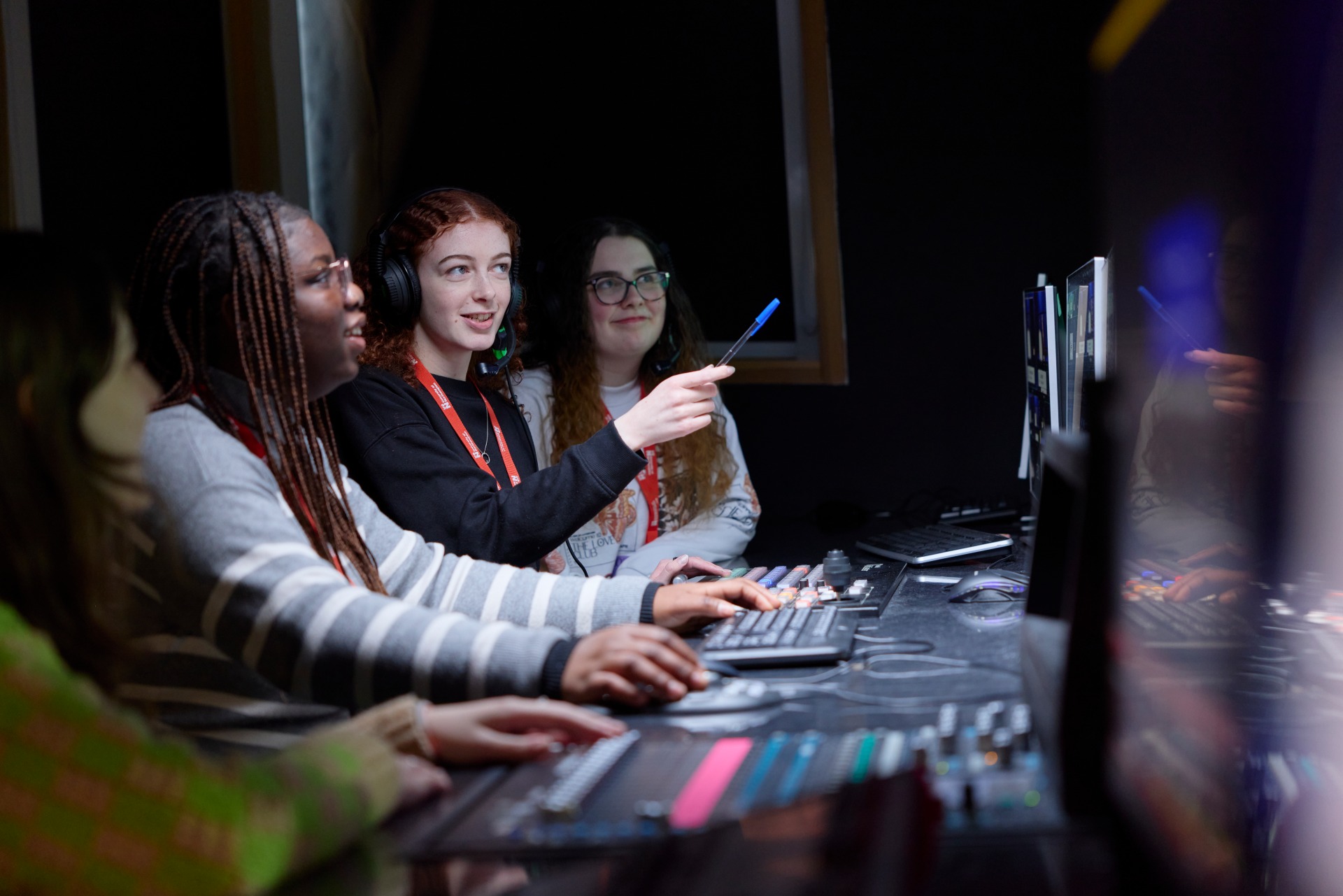 Student operating a radio desk with a microphone and headphones during a university journalism workshop, while another student takes notes in the foreground