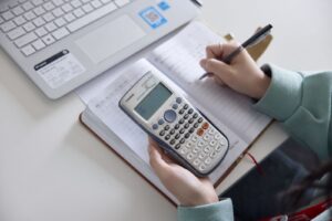 A student writes in a notebook while holding a scientific calculator, with a laptop open on the desk nearby.