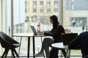 A person sits at a round table near large windows, working on a laptop with a water bottle beside them in a modern indoor setting.