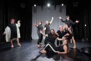 Eight students in black clothing pose energetically on a stage with black curtains and bright overhead lights; one person stands apart wearing a long white fluffy cardigan.