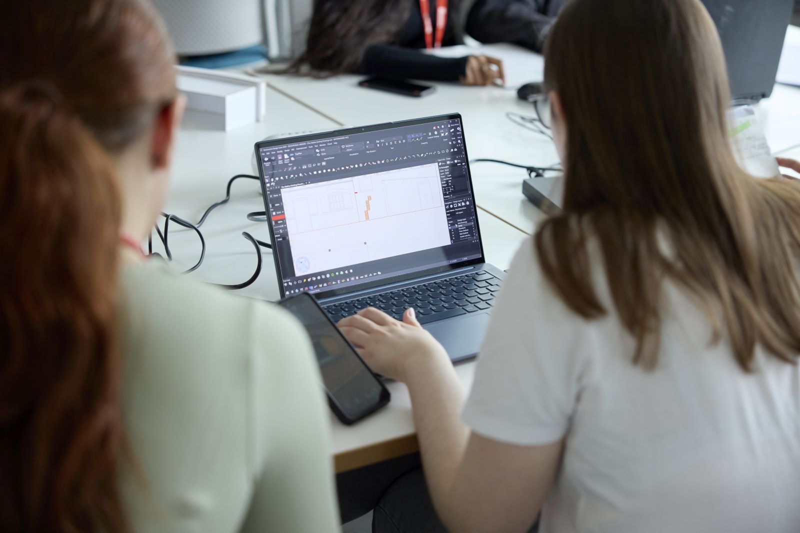 Two people work at a desk, looking at a laptop screen displaying a software interface with a technical or design drawing.