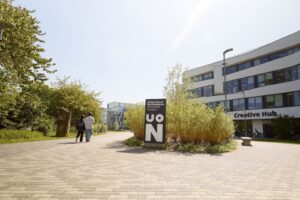 A paved walkway leads to a sign reading "University of Northampton Waterside Campus UON," with the Creative Hub building visible on the right. Two people walk along the path.