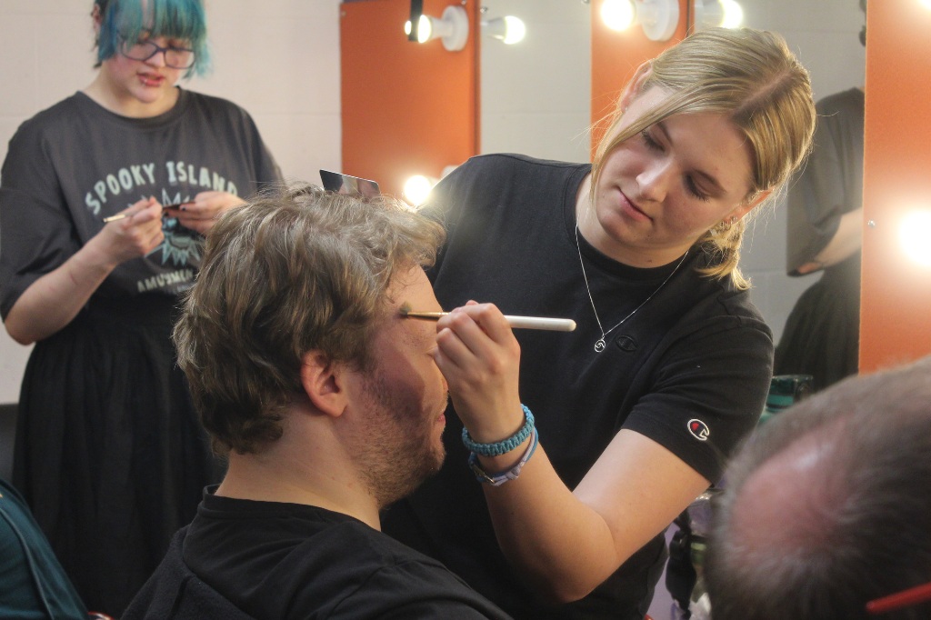 A woman applies makeup to a seated man while another person prepares items in the background near illuminated mirrors.