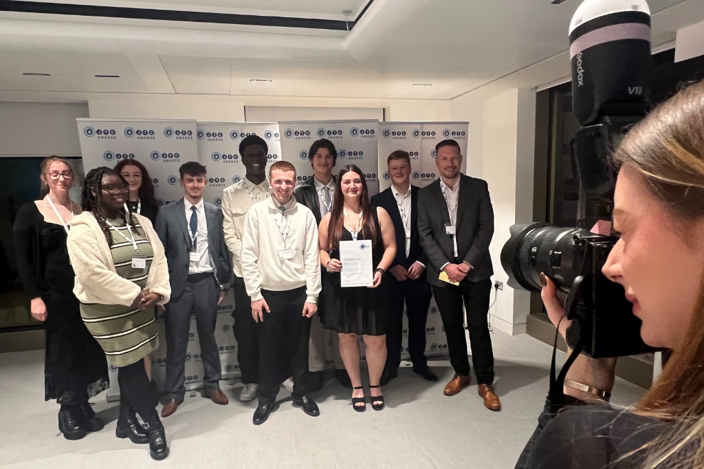A group of people pose for a photo, with one person holding a certificate, while a photographer takes their picture in an indoor setting with a branded backdrop.