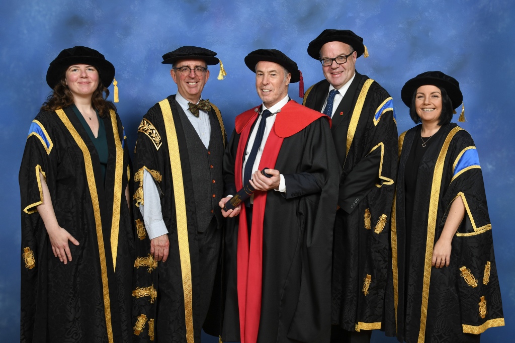 Five people pose in academic robes against a blue background; one person in the center wears red and black, holding a diploma, while the others wear black and gold regalia.