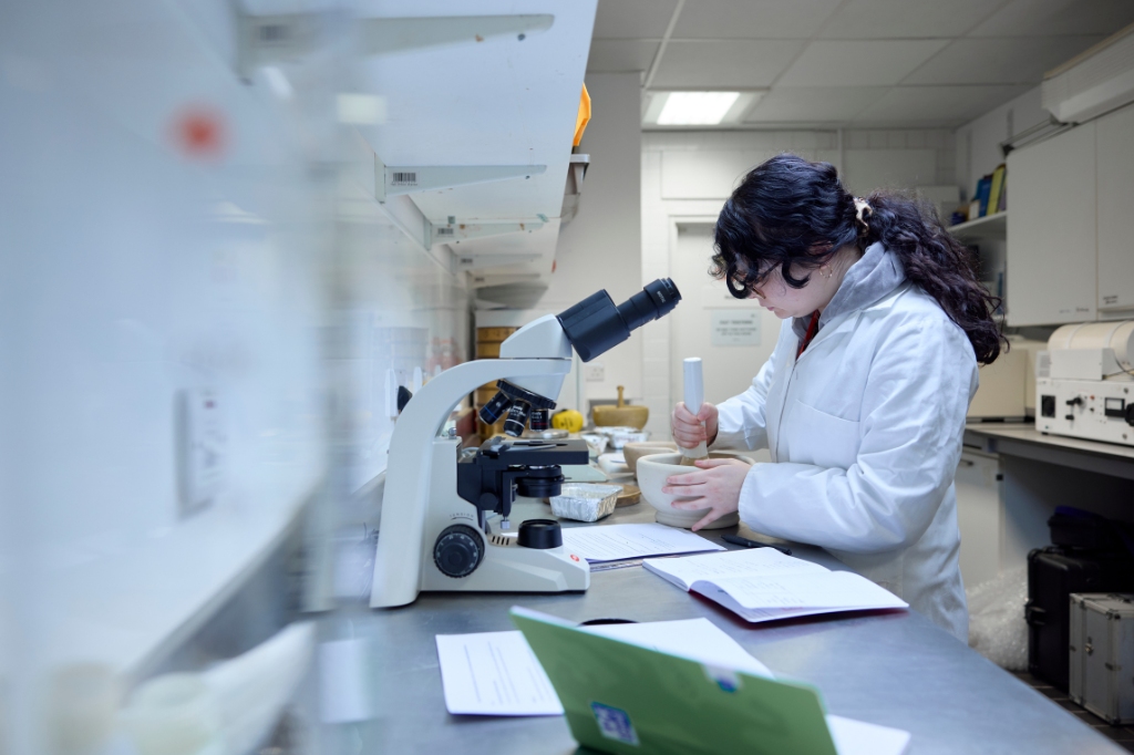 A person in a lab coat works at a laboratory bench, using a mortar and pestle near a microscope and open notebook.