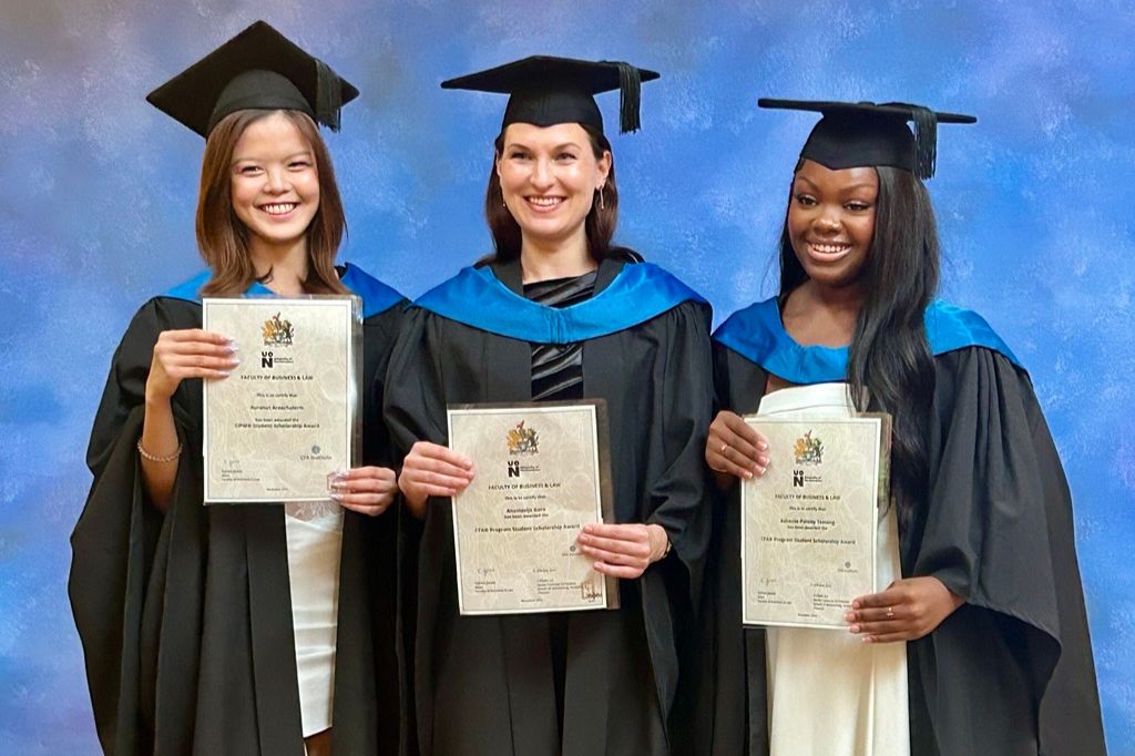 Three graduates - Ashanté-Paisley Temeng, Anastasija Gore, and Auranut Areechalerm - in caps and gowns stand side by side, smiling and holding certificates against a blue backdrop.