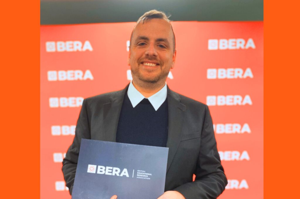 A person in a suit holds a BERA certificate in front of a red BERA-branded backdrop.