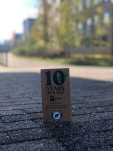 A wooden plaque commemorating 10 years (2015–2025) of BREEAM Green Accreditation stands on a paved outdoor path with blurred buildings and trees in the background.