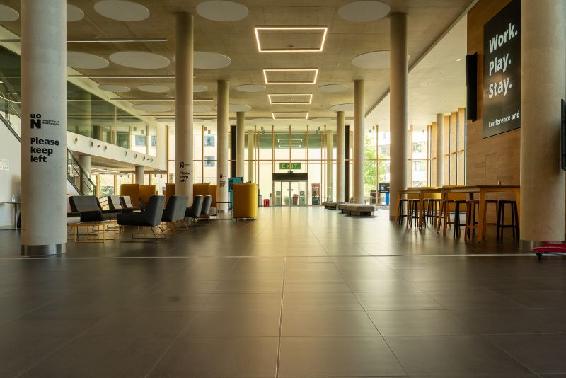 Spacious modern lobby with tiled floor, seating areas, tall columns, large windows, and natural light. Signs and digital screens are visible on the walls.