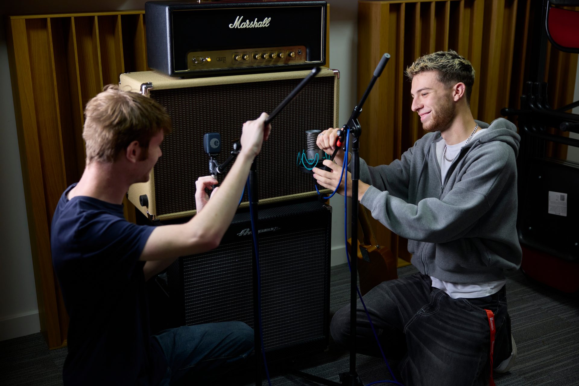 Two students kneeling on the floor in a recording studio, adjusting microphones and cables in front of a large Marshall amplifier stack. Both are focused on setting up the audio equipment, with wooden acoustic panels in the background.
