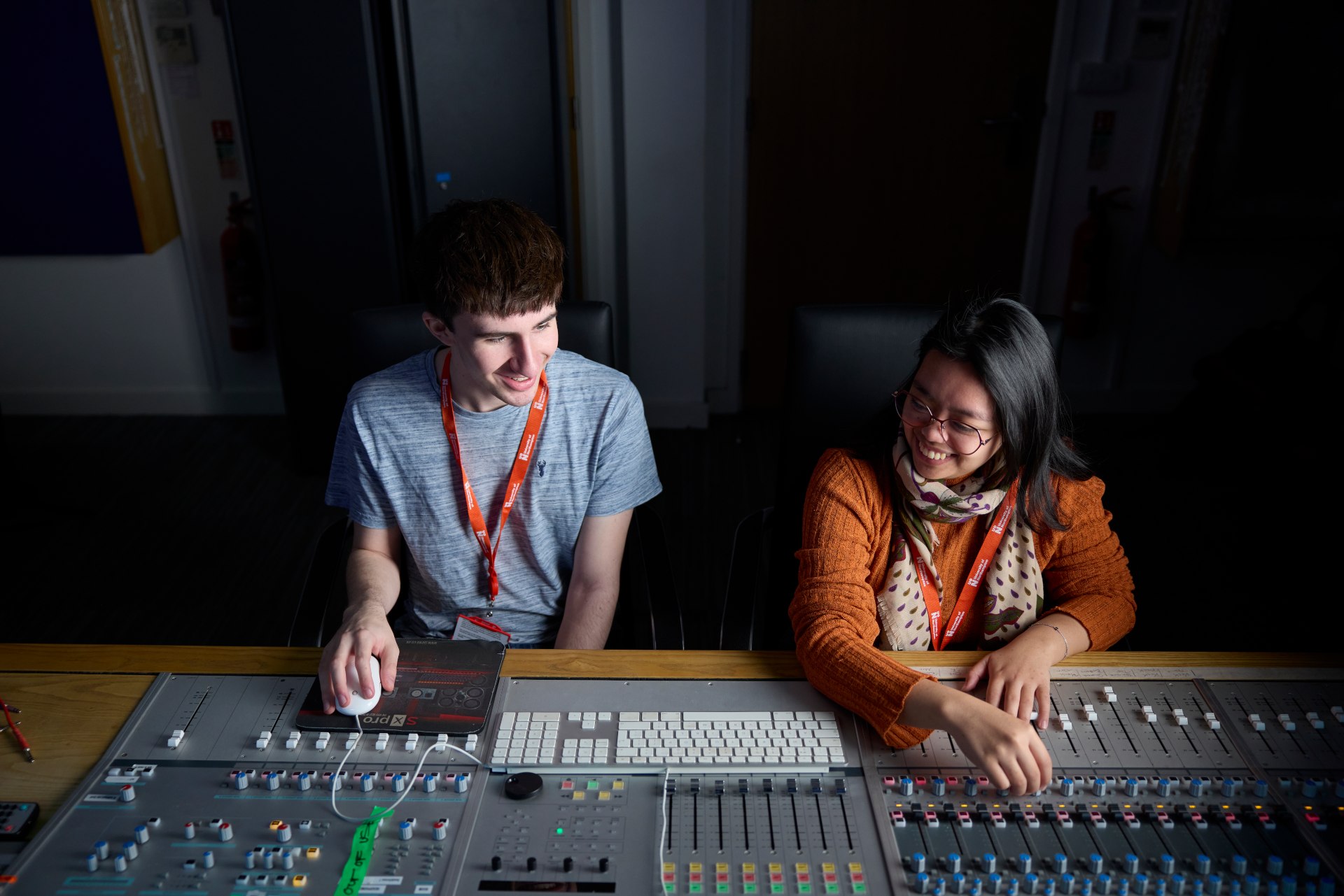 Two students working at a professional audio mixing console in a recording studio, adjusting controls and monitoring equipment