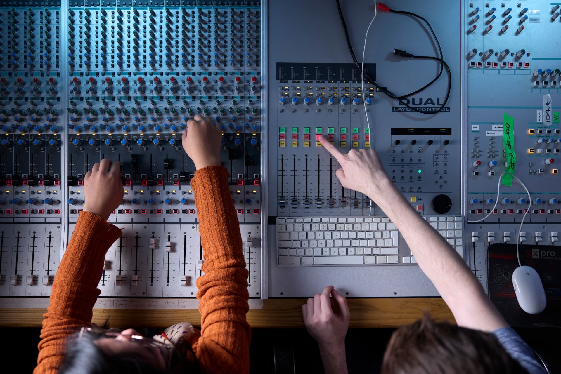 Top-down view of two students operating an audio mixing desk, with one person adjusting multiple knobs and sliders while the other points to illuminated buttons on a digital control panel. The console is covered with colourful controls, cables, and a computer keyboard.