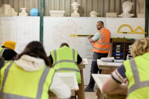 A tutor with a spirit level in hand wearing an orange his vis vest teaching a class of students in green vests.