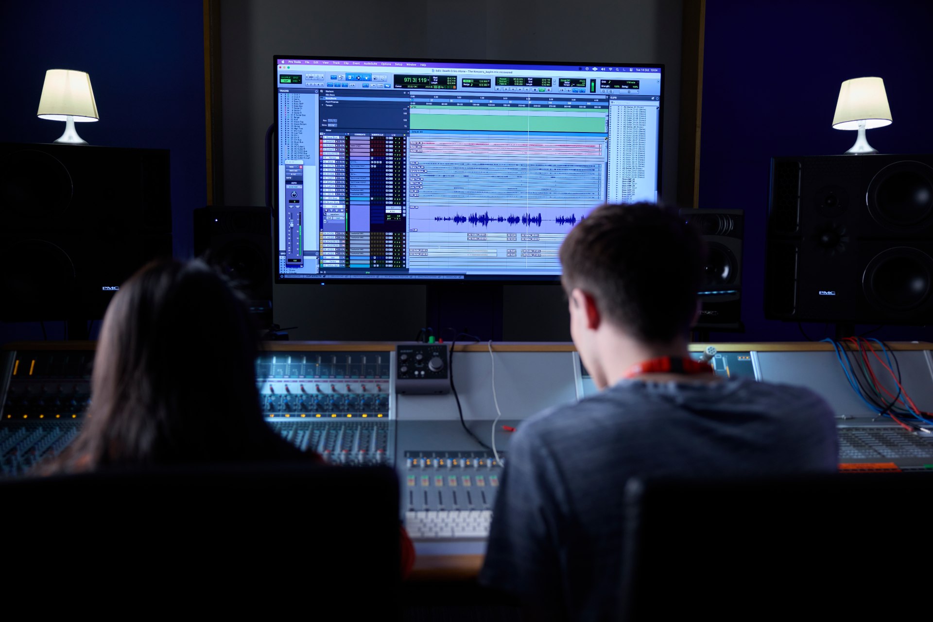 Two students seated at a professional audio mixing console in a recording studio, facing a large monitor displaying digital audio editing software with multiple tracks and waveforms. The setup includes speakers on both sides and two small lamps in the background.